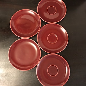 May include: Five maroon-colored ceramic saucers and plates. The saucers have a central recessed area for a cup. The plates are solid maroon. The items are arranged on a dark surface.