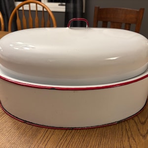 May include: White enamel roasting pan with a matching lid. The pan and lid have red trim and handles. The lid has a red handle. The pan is sitting on a wooden table.