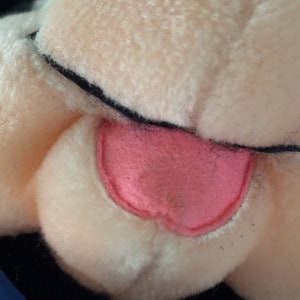 May include: Close-up of a plush toy's face, featuring a pink tongue and white fur. The toy has black eyes and a black nose.