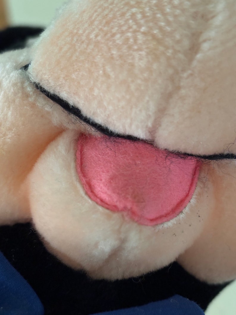 May include: Close-up of a plush toy's face, featuring a pink tongue and white fur. The toy has black eyes and a black nose.
