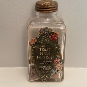 May include: A clear glass bottle with a gold-colored lid. The bottle has a decorative label with the text "TOUT MON JARDIN" and floral accents. The label also includes the text "POT-POURRI SACHET" and "RICHARD HUDNUT NEW-YORK PARIS".
