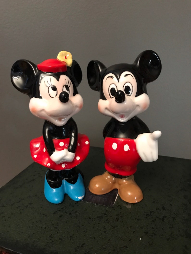Pu&ograve; includere: Un set di figurine in ceramica vintage di Topolino e Minnie Mouse. Topolino indossa pantaloncini rossi con pois bianchi e scarpe marroni. Minnie indossa un vestito rosso con pois bianchi, un cappello rosso con un fiore giallo e scarpe blu.
