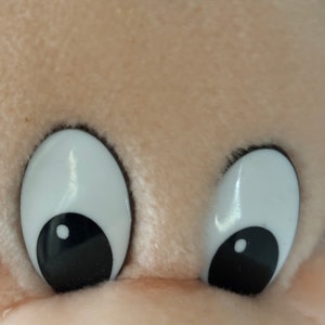 May include: Close-up of a plush toy's face with large white eyes and black pupils. The toy has a light brown fur texture.