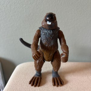 May include: A brown plastic action figure of a beaver with a gray chest and gray wrist and ankle bands. The beaver has a black nose, white teeth, and a small tail. The figure is standing on a beige surface.