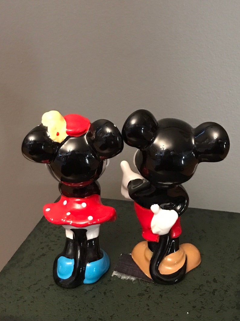 Pu&ograve; includere: Un set di figurine in ceramica di Topolino e Minnie Mouse. Topolino indossa pantaloncini rossi e scarpe nere. Minnie indossa un vestito a pois rossi e bianchi e scarpe blu. Entrambe le figurine hanno orecchie nere e occhi neri.
