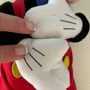 May include: A plush Mickey Mouse toy with red shorts, yellow buttons, and white gloves. The toy is in a standing position with its arms raised.
