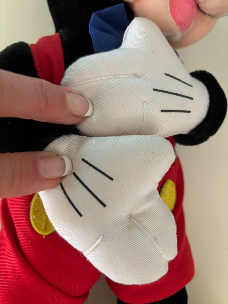 May include: A plush Mickey Mouse toy with red shorts, yellow buttons, and white gloves. The toy is in a standing position with its arms raised.
