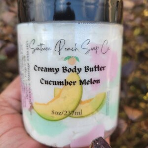 Może przedstawiać: Biały słoik z czarną pokrywką zawierający Krem do Ciała Creamy Body Butter Cucumber Melon firmy Southern Peach Soap Co. Słoik jest oznaczony zielonym i żółtym plastrkiem melona oraz tekstem "8oz/237ml".