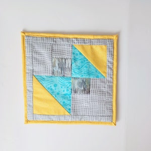May include: A yellow and gray quilted coaster with a blue and white geometric pattern in the center.