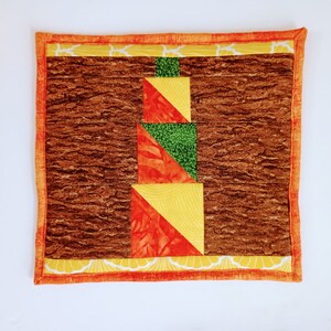 May include: A brown quilted coaster with a geometric design in orange, yellow, and green. The coaster has a decorative border with a floral pattern.