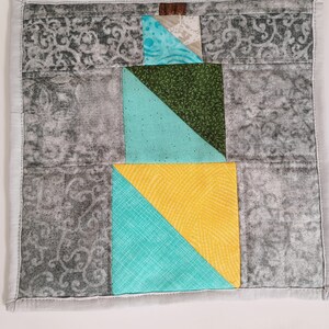 Può includere: Un quadrato di tessuto grigio e bianco con un design a patchwork. Il patchwork include un tessuto blu turchese, giallo, verde e marrone. Il tessuto è cucito con un filo bianco.