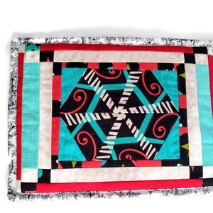 May include: A colourful patchwork quilt with a geometric design in shades of red, white, black, and turquoise. The quilt is framed with a white border and a black and white patterned border.