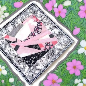 May include: A pink and black patchwork quilt square with a white background and black and white trim. The square is surrounded by a white background with a gray border.