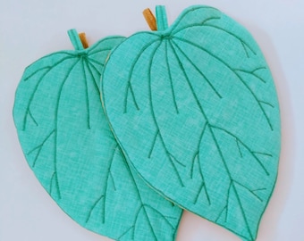 May include: Two teal green leaf-shaped pot holders with a brown fabric trim. The pot holders are quilted and have a detailed embroidered leaf design.