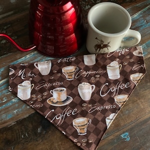 May include: A brown bandana with a checkered pattern and coffee-themed illustrations. The bandana features various coffee drinks, including cappuccino, latte, mocha, and espresso, along with coffee beans and the word "Coffee".
