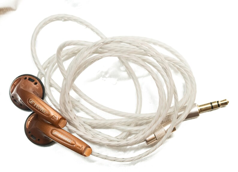 K&ouml;nnte beinhalten: Ein Paar kupferfarbene In-Ear-Kopfh&ouml;rer mit einem wei&szlig;en geflochtenen Kabel und einem vergoldeten 3,5-mm-Stecker.