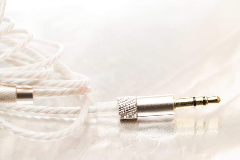 K&ouml;nnte beinhalten: Nahaufnahme eines wei&szlig;en geflochtenen Audiokabels mit einem vergoldeten 3,5-mm-Klinkenstecker.