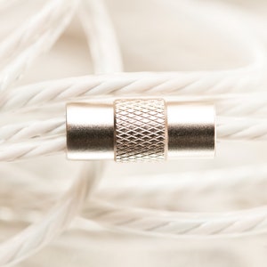 K&ouml;nnte beinhalten: Nahaufnahme eines silbernen Steckers mit strukturiertem Design, umgeben von einem wei&szlig;en geflochtenen Kabel.