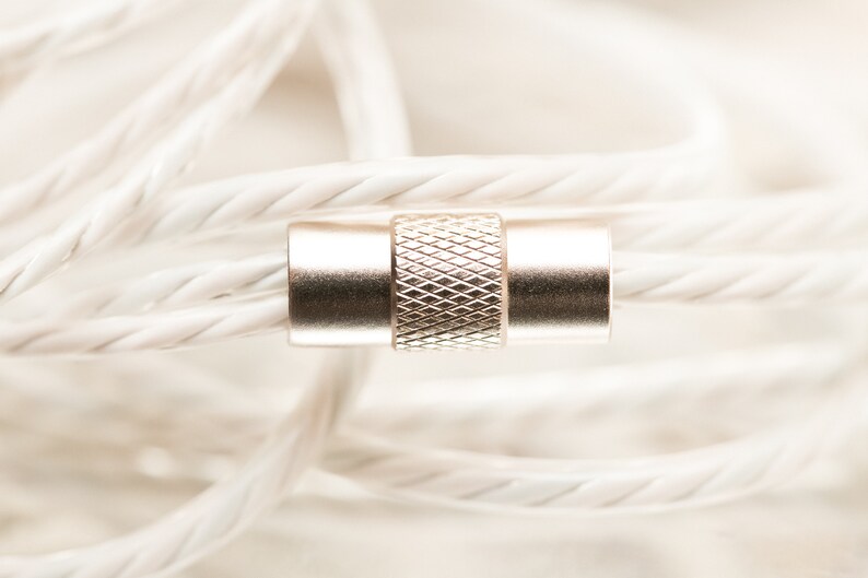 K&ouml;nnte beinhalten: Nahaufnahme eines silbernen Steckers mit strukturiertem Design, umgeben von einem wei&szlig;en geflochtenen Kabel.