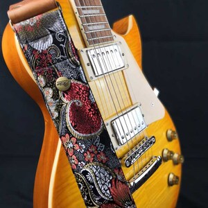 May include: A close-up of a sunburst electric guitar with a decorative guitar strap. The strap features a black background with red and white floral embroidery and a brown leather end. The guitar has chrome hardware.