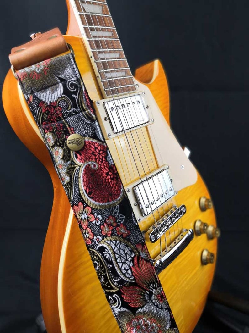 May include: A close-up of a sunburst electric guitar with a decorative guitar strap. The strap features a black background with red and white floral embroidery and a brown leather end. The guitar has chrome hardware.