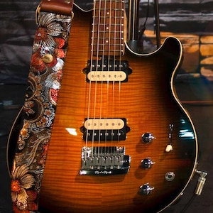 May include: A sunburst-finish electric guitar with a floral patterned strap. The guitar has a brown and orange finish, chrome hardware, and six strings. The strap features a colorful floral design with red, orange, and silver accents.