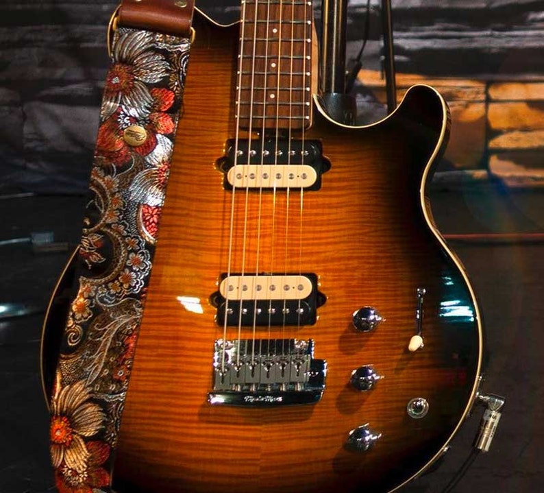 May include: A sunburst-finish electric guitar with a floral patterned strap. The guitar has a brown and orange finish, chrome hardware, and six strings. The strap features a colorful floral design with red, orange, and silver accents.