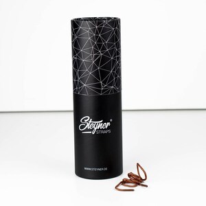 May include: A black cylindrical box with a geometric pattern on the lid and the "Steyner STRAPS" logo in white. A pair of brown shoelaces is next to the box. The website address www.steyner.de is printed on the box.
