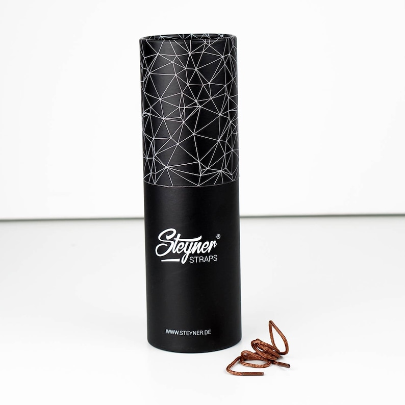 May include: A black cylindrical box with a geometric pattern on the lid and the "Steyner STRAPS" logo in white. A pair of brown shoelaces is next to the box. The website address www.steyner.de is printed on the box.