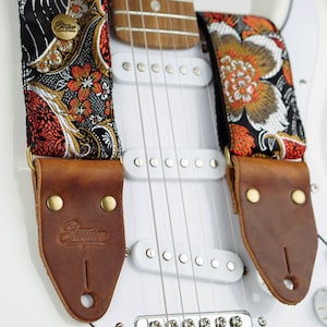 May include: A guitar strap with a floral pattern in orange, gold, and white on a black background. The strap has brown leather ends with the brand name "Stegner" embossed. The strap is attached to a white electric guitar.