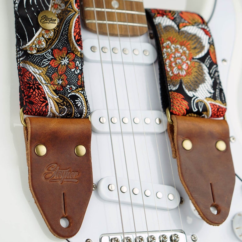May include: A guitar strap with a floral pattern in orange, gold, and white on a black background. The strap has brown leather ends with the brand name "Stegner" embossed. The strap is attached to a white electric guitar.