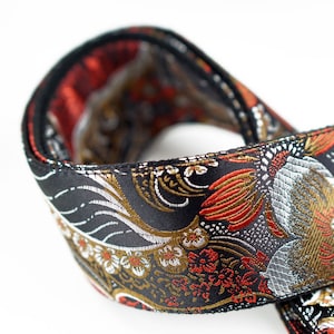 May include: Close-up of a black strap with an ornate floral and bird pattern. The design features red, gold, and silver accents on a black background. The strap is folded over, revealing a red interior.