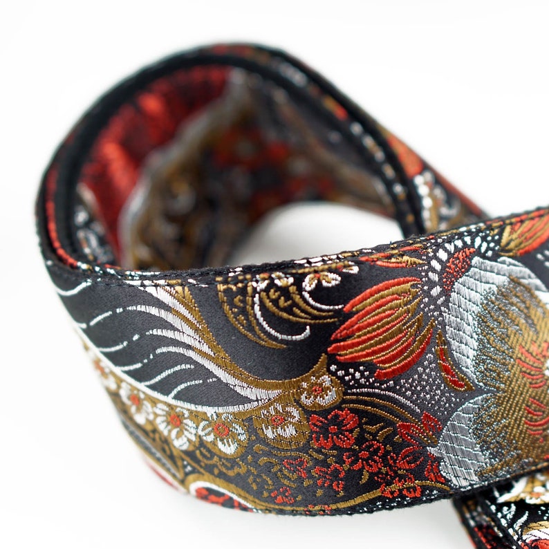 May include: Close-up of a black strap with an ornate floral and bird pattern. The design features red, gold, and silver accents on a black background. The strap is folded over, revealing a red interior.