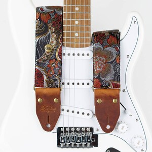 May include: A guitar strap with a floral pattern in red, gold, and silver on a black background. The strap has brown leather ends with gold rivets and the brand name "Stegler" embossed on the leather. The strap is attached to a white electric guitar.