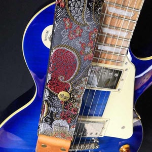 May include: A decorative guitar strap with a floral pattern in red, silver, and black. The strap is attached to a blue electric guitar. The strap has a brown leather end piece and a gold-colored metal buckle.