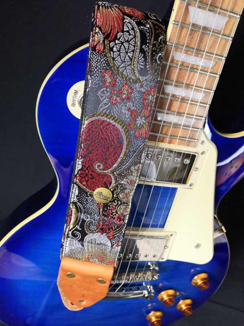May include: A decorative guitar strap with a floral pattern in red, silver, and black. The strap is attached to a blue electric guitar. The strap has a brown leather end piece and a gold-colored metal buckle.