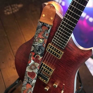May include: A reddish-brown electric guitar with a floral patterned strap. The strap features a black background with red, silver, and orange flowers. The guitar has gold hardware and a wooden body.