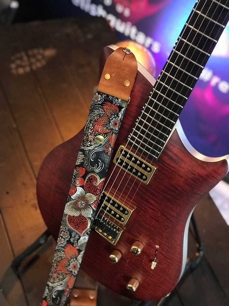 May include: A reddish-brown electric guitar with a floral patterned strap. The strap features a black background with red, silver, and orange flowers. The guitar has gold hardware and a wooden body.