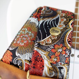 May include: Close-up of a guitar strap with a floral pattern in black, gold, silver, and red. The strap has a brown leather backing and a brass button with the word "Steyner" on it. The strap is attached to a white guitar.