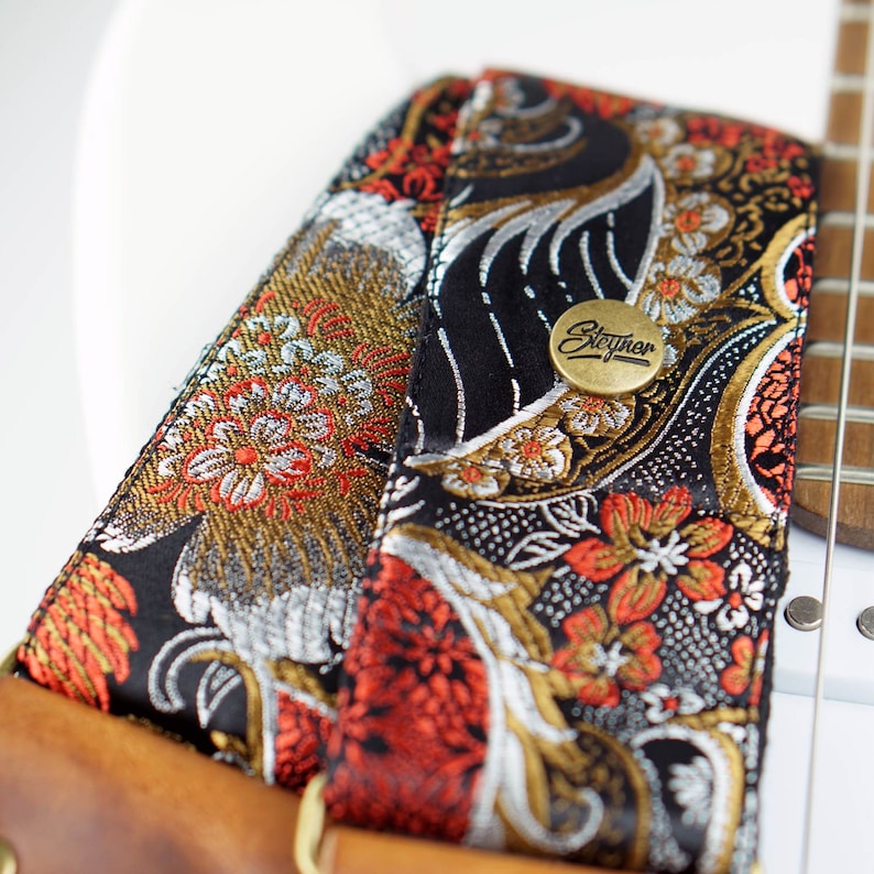 May include: Close-up of a guitar strap with a floral pattern in black, gold, silver, and red. The strap has a brown leather backing and a brass button with the word "Steyner" on it. The strap is attached to a white guitar.