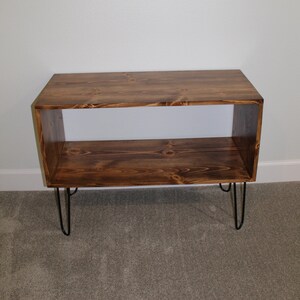 May include: A wooden shelf unit with two shelves and black hairpin legs. The shelf is made of dark brown wood and has a natural wood grain.