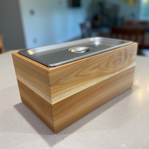 May include: A wooden box with a stainless steel insert. The box is made of light wood with a darker wood trim. The box is sitting on a light gray surface.