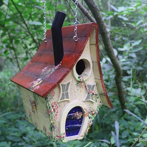 May include: A whimsical birdhouse painted in shades of red, white, and blue, with a chimney and a blue door. The birdhouse is decorated with floral details and has a small opening for birds to enter.