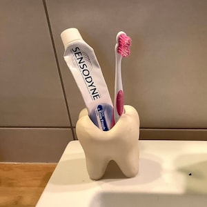 Peut inclure: Un porte-brosse à dents en céramique blanc en forme de dent, contenant un tube de dentifrice Sensodyne et une brosse à dents rose et blanche. Le support est sur un évier blanc à côté d'un robinet chromé.