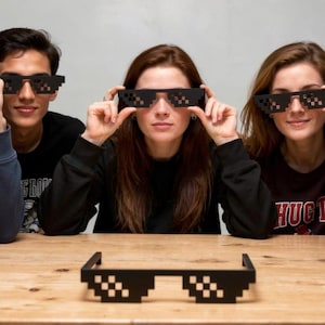 Peut inclure: Cinq personnes tenant des lunettes de soleil noires pixélisées, avec une paire de ces mêmes lunettes posées sur une table en bois. Les lunettes de soleil ont un design pixélisé distinctif, créant une esthétique rétro.