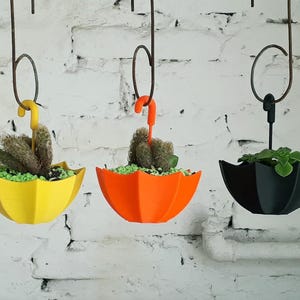 Peut inclure: Trois jardinières suspendues en forme de parapluie, jaunes, oranges et noires. Chaque jardinière contient une petite plante et des cailloux verts décoratifs. Les jardinières sont suspendues à des crochets métalliques contre un mur de briques blanches.