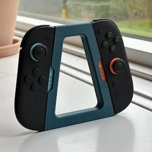 Peut inclure: Une manette Nintendo Switch noire et sarcelle avec un cadre sarcelle. La manette a des boutons noirs, un joystick bleu et un joystick orange. La manette repose sur une surface blanche.