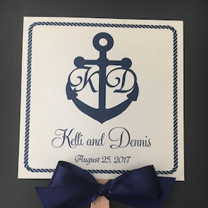 Puede incluir: Un abanico de boda blanco con un ancla azul marino y las iniciales "K" y "D" entrelazadas. El abanico tiene un lazo azul marino y el texto "Kelli y Dennis August 25, 2017".