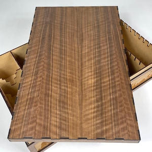 May include: A wooden box with a lid that opens to reveal compartments for organizing game pieces. The lid is made of a dark wood with a natural grain pattern.