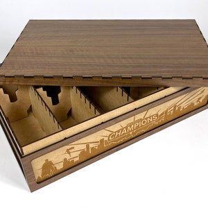 May include: A wooden box with a lid, designed to store and organize a board game. The box has a brown wood finish and features a laser-engraved design of the Marvel Champions logo and a cityscape. The interior of the box is divided into compartments for storing game components.